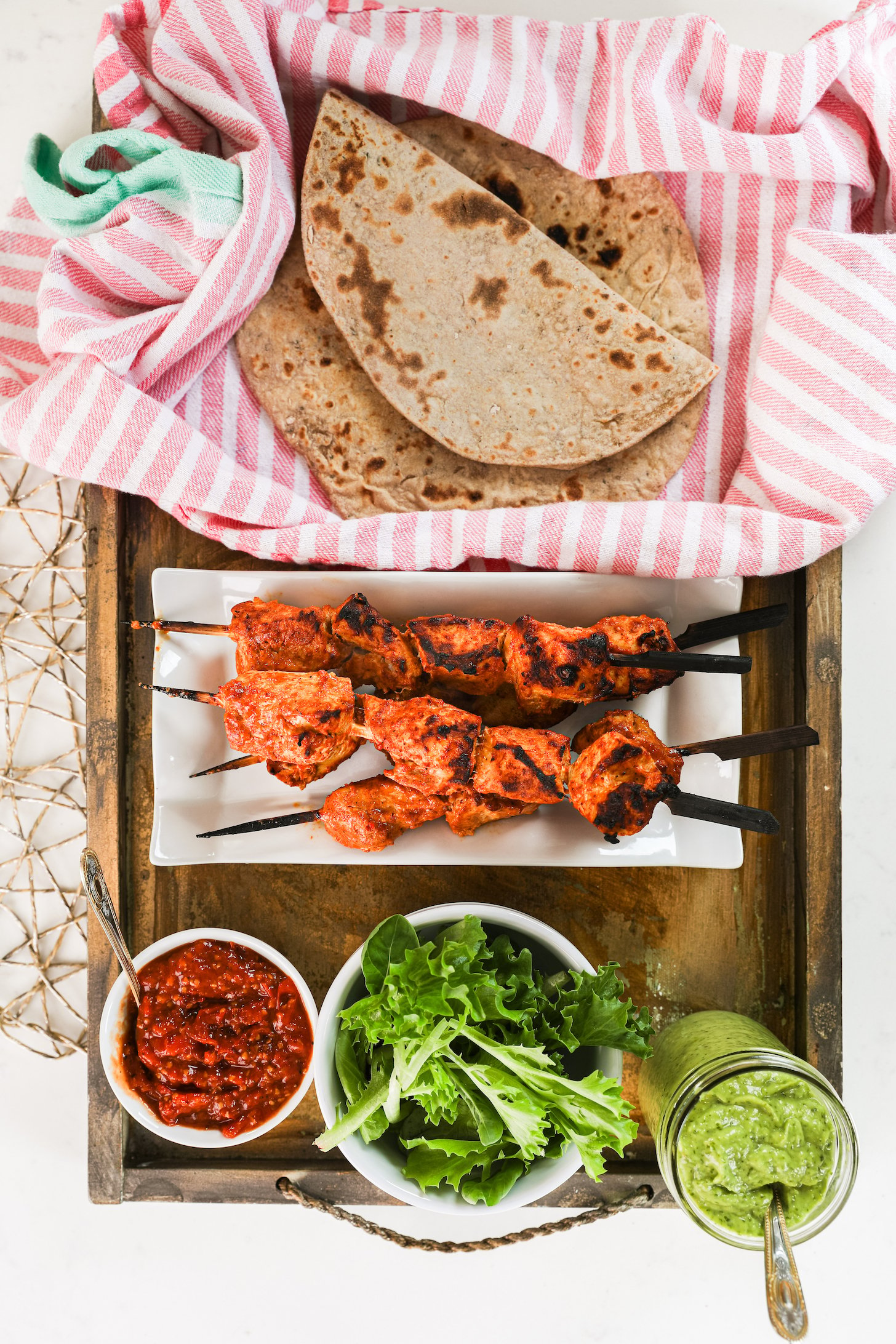 A selection of food ingredients like grilled tandoori chicken skewers, greens, sauces and roti.