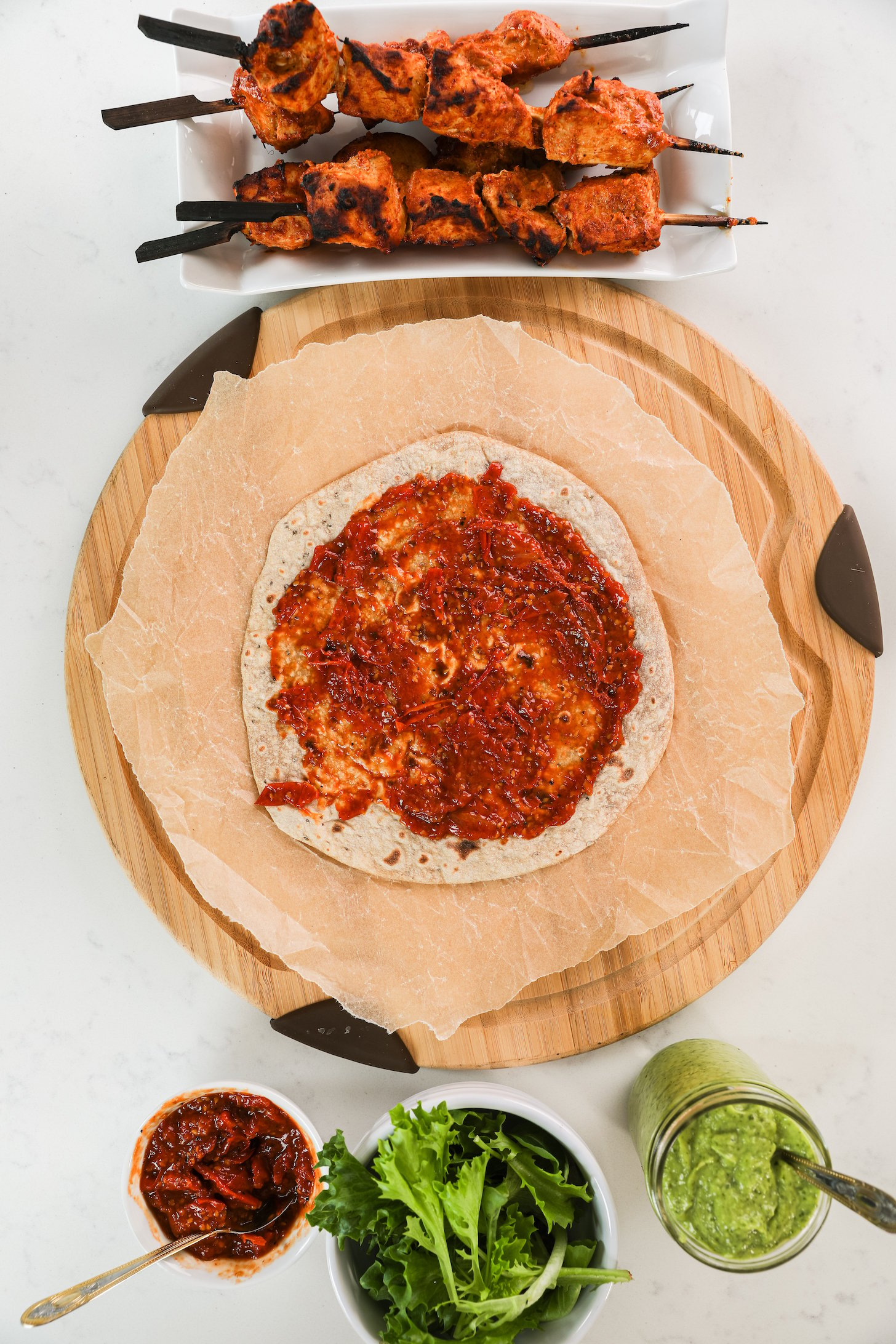 A roti spread with tomato sauce with tandoori chicken skewers, sauces and salad greens nearby.