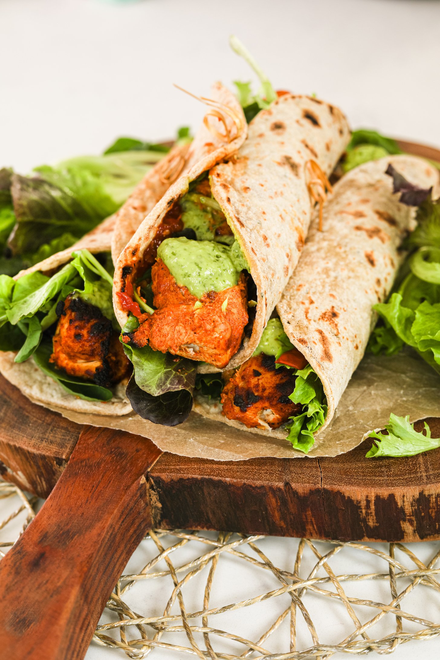 Chicken tikka wraps piled on top of one another surrounded by greens on a wooden board.