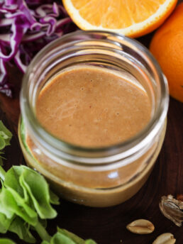 A close up perspective image of a small jar of dressing surrounded by an orange half, peanuts.