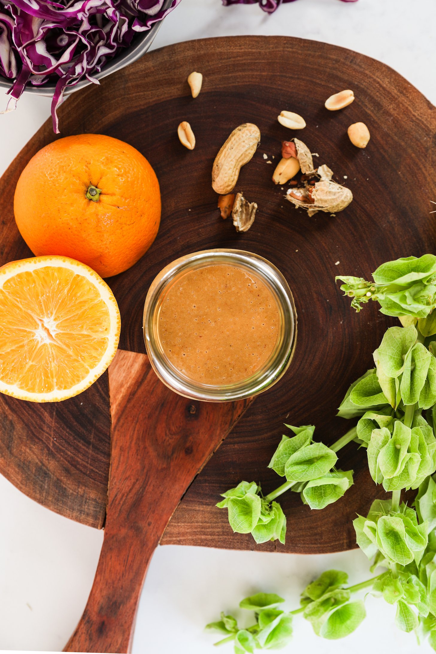 Flat lay of a small jar of dressing surrounded by an orange half, peanuts, and a green plant.