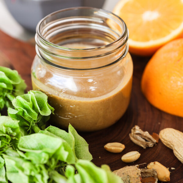 A perspective image of a small jar of dressing surrounded by an orange half, peanuts, sliced cabbage in the background and a green plant in the foreground.