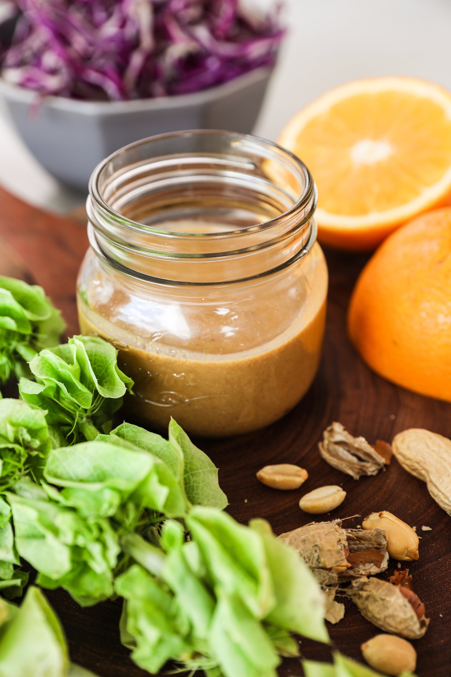 A perspective image of a small jar of dressing surrounded by an orange half, peanuts, sliced cabbage in the background and a green plant in the foreground.