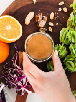 A hand holding a small jar of dressing surrounded by an orange half, peanuts, sliced cabbage and a green plant.