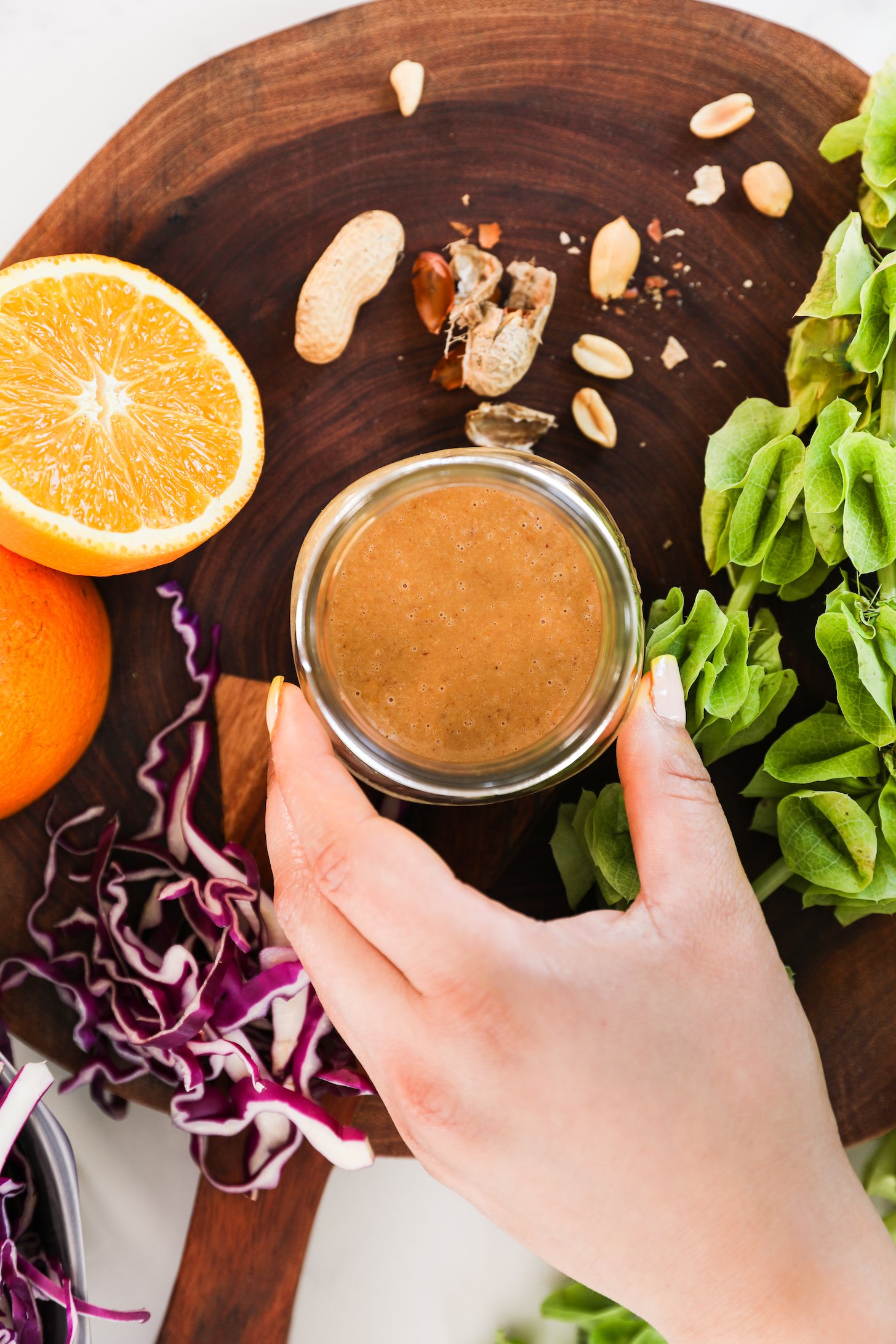 A hand holding a small jar of dressing surrounded by an orange half, peanuts, sliced cabbage and a green plant.