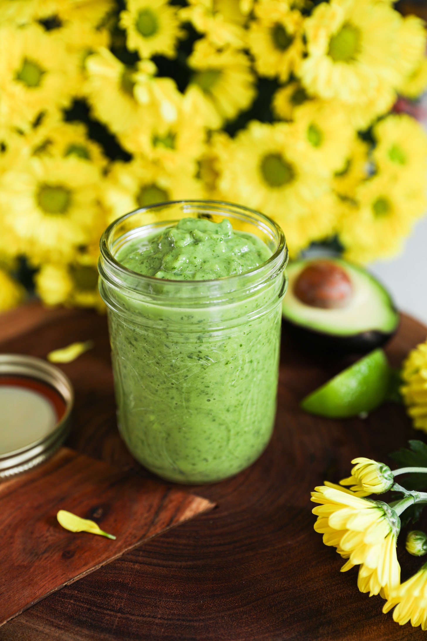 Side view of a mason jar filled with the avocado dressing with a bunch of yellow flowers in the background.
