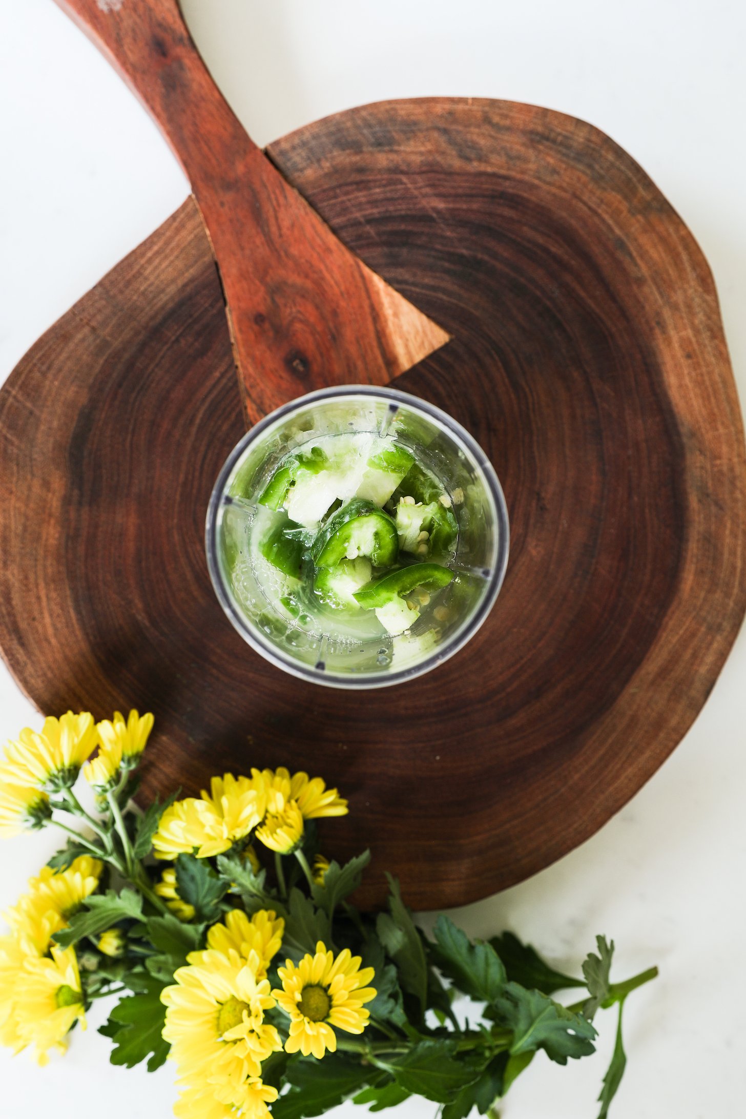 Top view of a small blender filled with chopped jalapeno and lime juice on a wooden board with yellow flowers nearby.