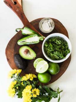 A selection of food ingredients like limes, avocado, jalapeno and herbs on a wooden board styled with yellow flowers.