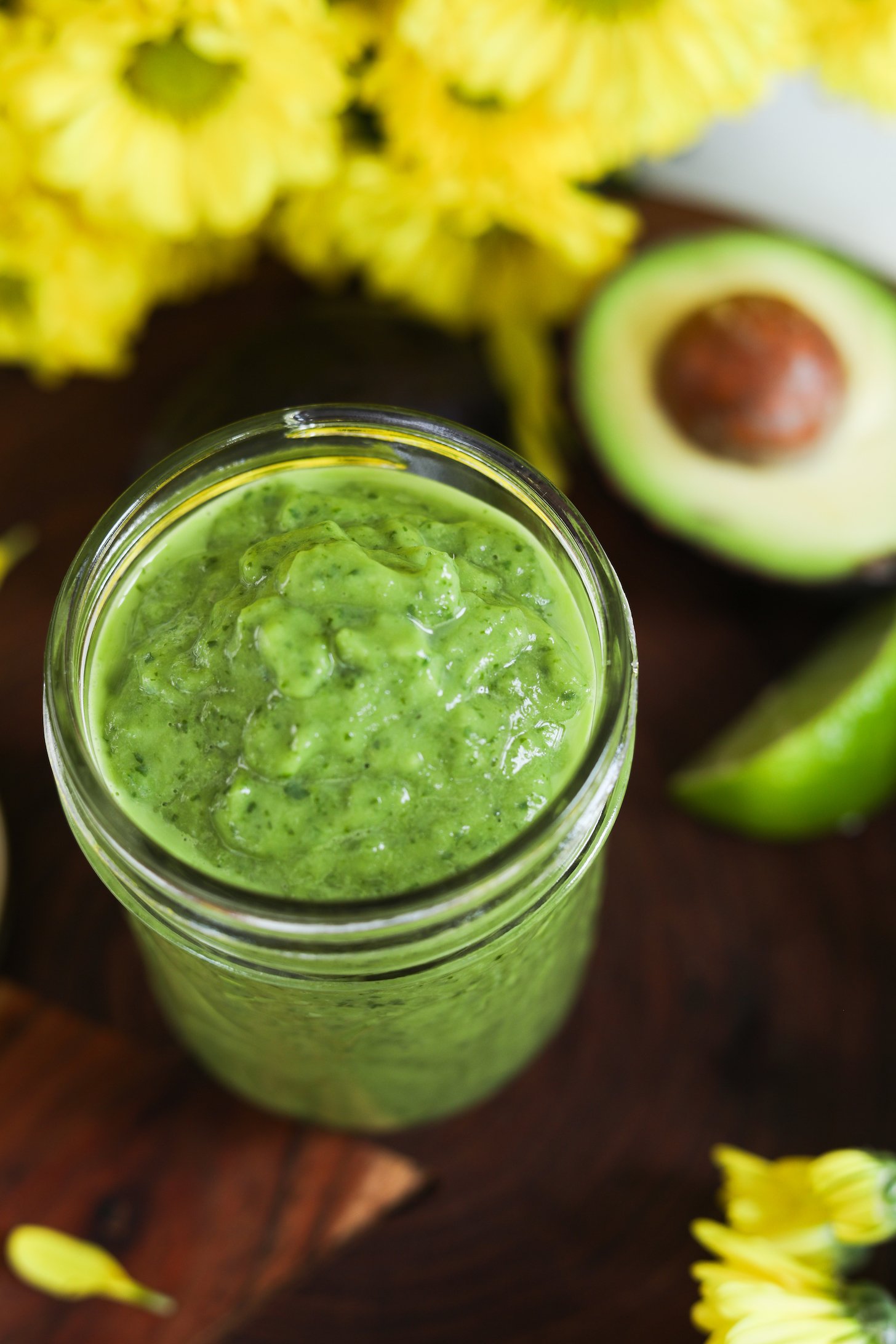 A jar of green creamy dressing with yellow flowers and avocado in the background.
