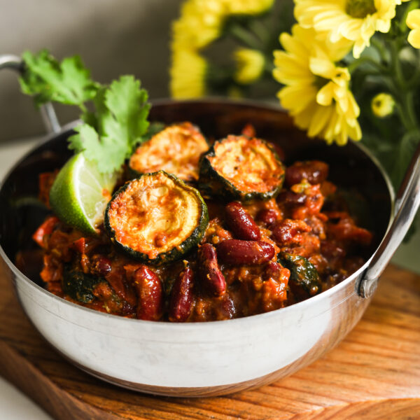 An Indian Karahi bowl of red kidney bean curry with zucchini styled with yellow flowers in the backgorund.