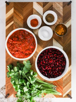 A selection of food ingredients like bowls of kidney beans, tomato sauce and ramekins of spices and herbs on a wooden board.