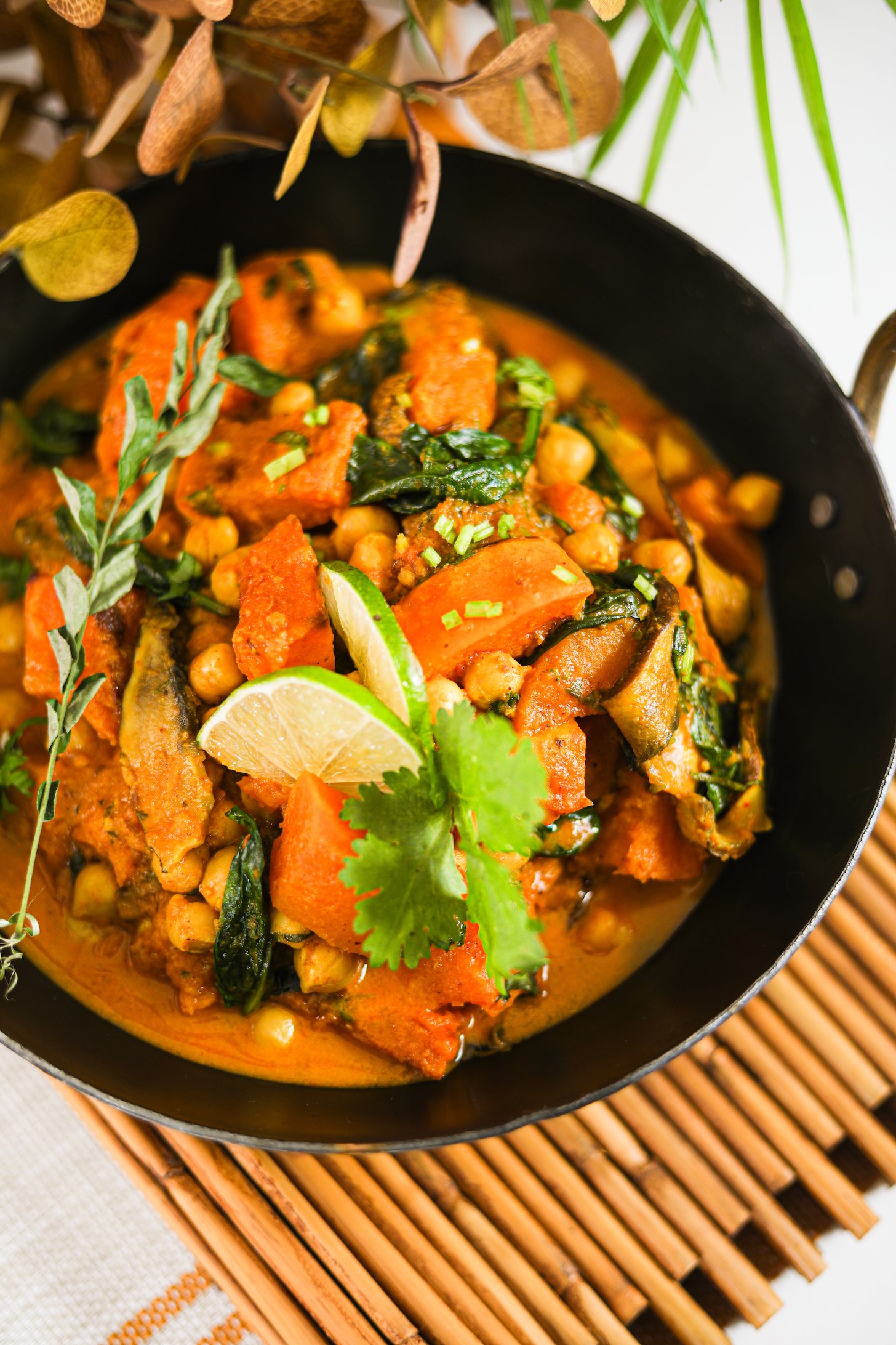 Perspective image of chickpea and squash curry in an Indian wok, topped with lime and cilantro on a bamboo mat.