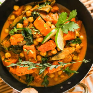 An Indian wok filled with chickpea and squash curry, garnished with lime slices and cilantro leaves on a bamboo mat.