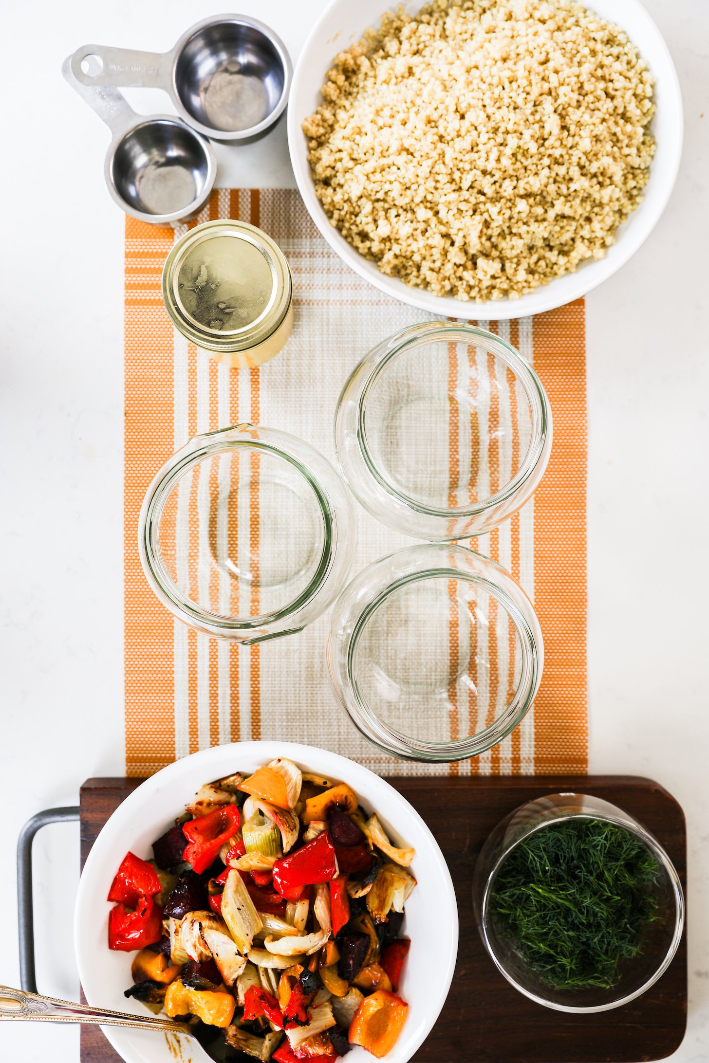 An arrangement featuring empty glass jars, a bowl filled with cooked quinoa, a vibrant selection of vegetables, and a bowl of freshly chopped dill—all set and ready for the assembly of a salad.