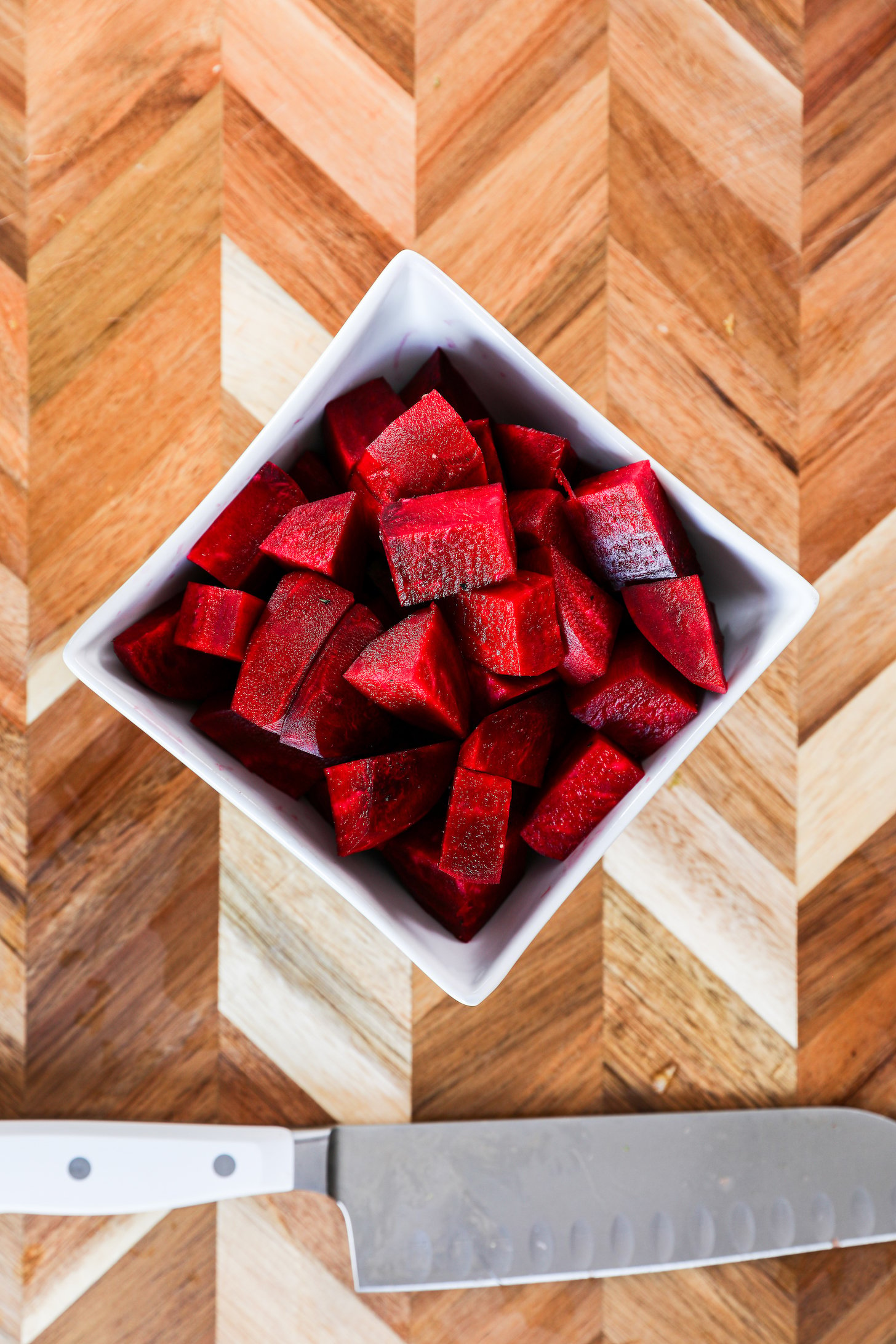 A bowl filled with cubed beetroots resting beside a knife on a wooden chopping board.