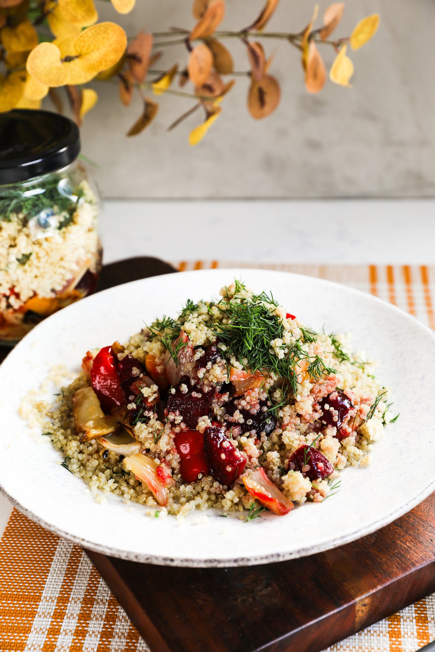 A plate of salad featuring a delightful mixture of quinoa, roasted beets, peppers, and fennel, topped with dill and doused in dressing with a glass salad jar nearby.