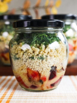 A close up of a glass jar of salad layered with dressing, veggies, quinoa and dill.