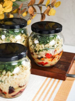 A dynamic perspective view of a vibrant display featuring three glass jars filled with salad layers, highlighting dressing, veggies, quinoa, and dill, set against a backdrop adorned by yellow leaves of a plant.