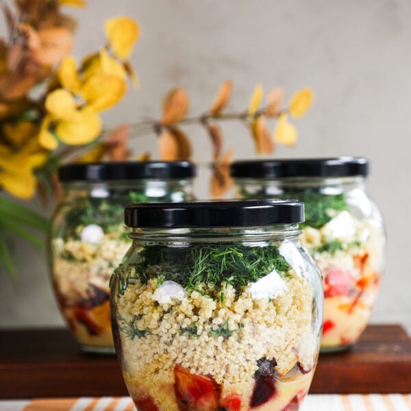 A vibrant display featuring three glass jars filled with salad layers, showcasing dressing, veggies, quinoa, and dill, with the backdrop adorned by yellow leaves of a plant.