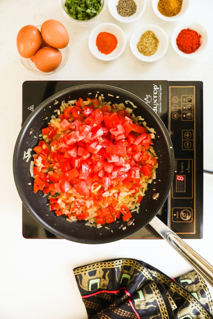 An image showing a cooking pan on a mobile stovetop filled with freshly chopped red peppers, tomatoes and onions, with ramekins of spices and a bowl of eggs placed nearby.