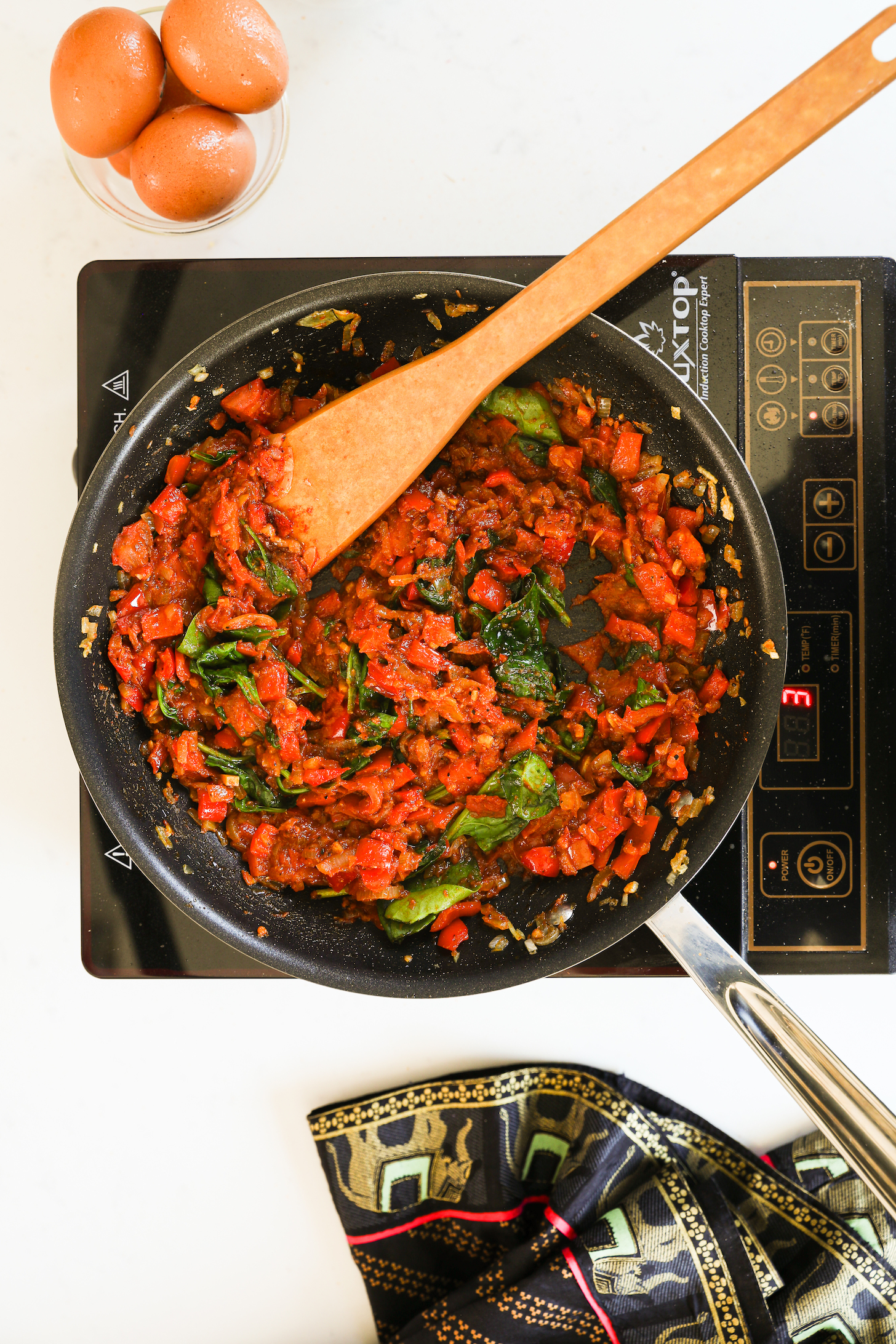 Image of a pan on a portable stove with cooked tomatoes peppers and greens inside. There is a bowl of eggs nearby.
