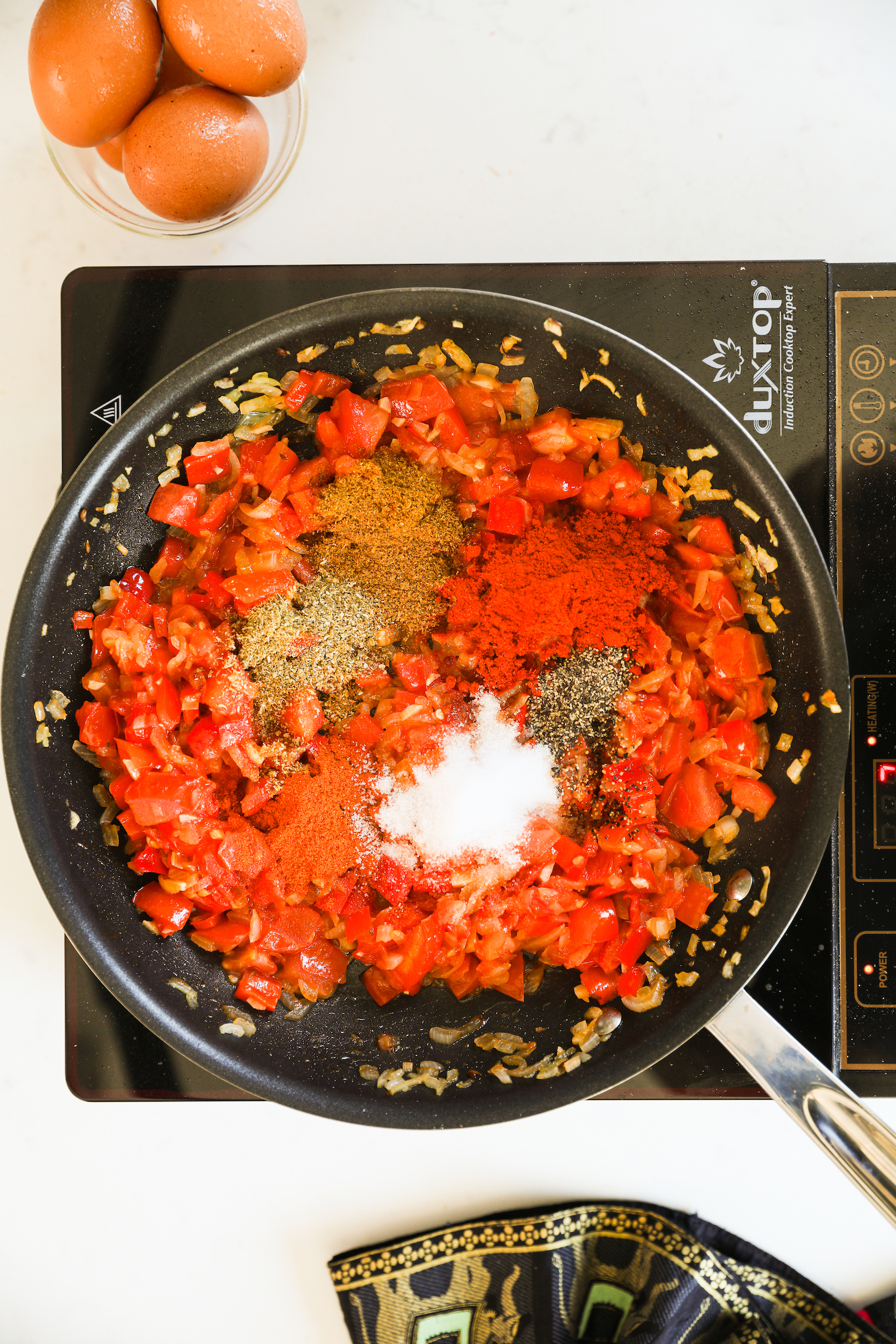 Image of a pan on a portable stove with cooked tomatoes and peppers inside. There are spices on top, and you can see a bowl of eggs nearby.