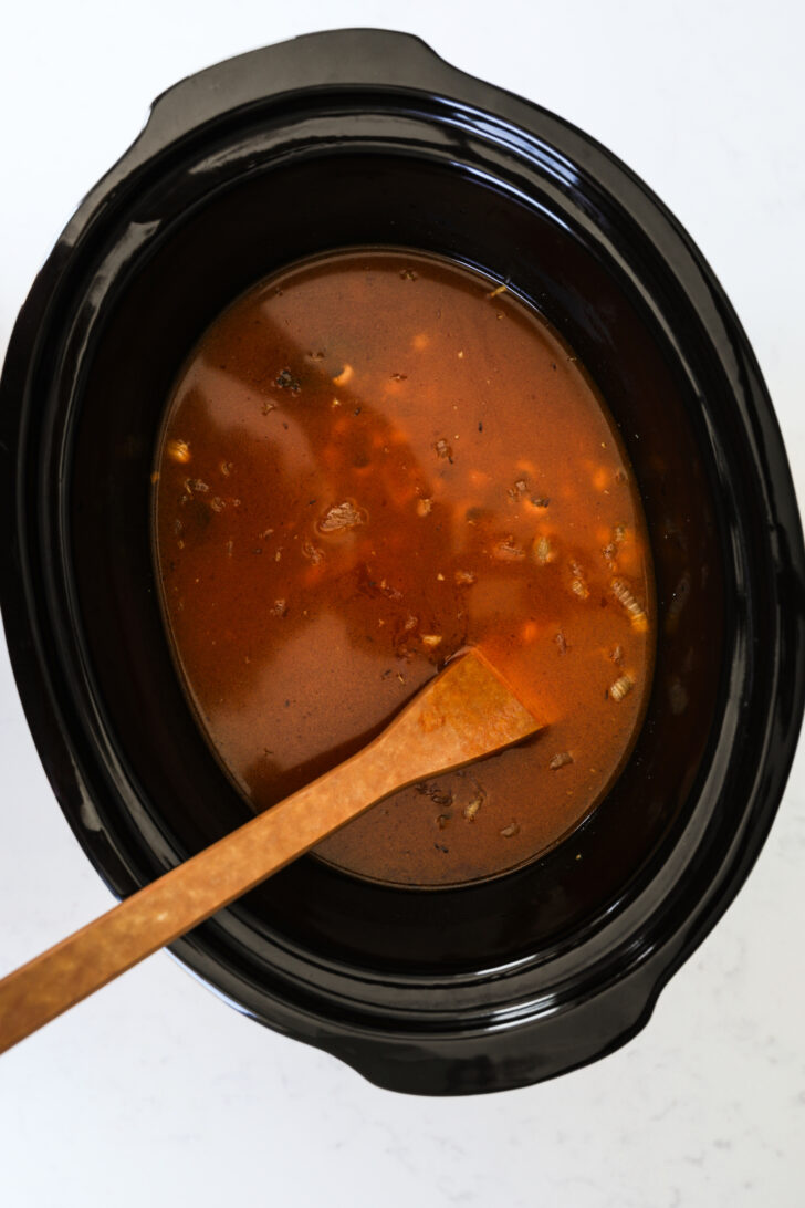 Flatlay image showcasing a slow cooker filled with black-eyed peas and completely immersed in a red broth, featuring a wooden utensil also submerged.