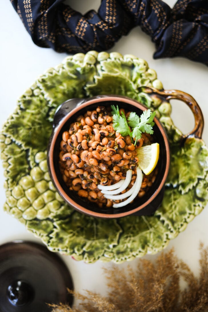 Flatlay image of a clay serving pot filled to the brim with black eyed peas, aka lobia curry topped with cilantro, lemon and onion slices.