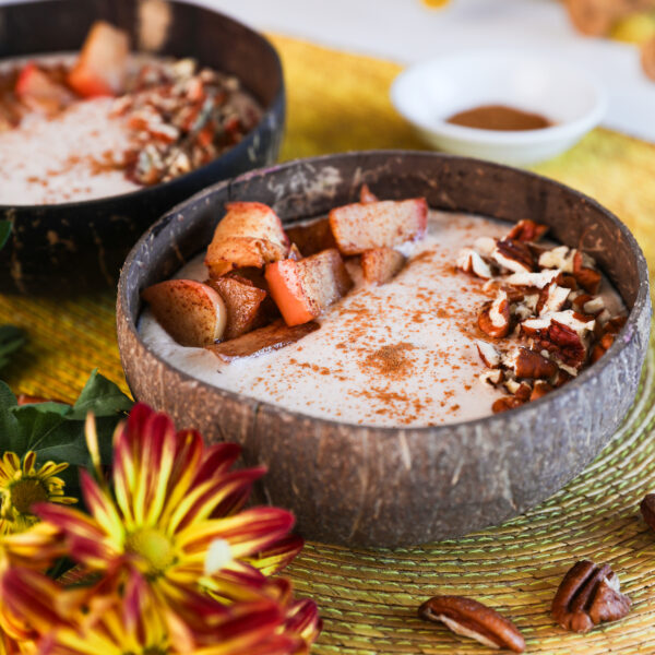 Image of two coconut bowls filled with beige smoothie, garnished with baked apple slices, pecans, and cinnamon, with bright yellow flowers in the foreground.