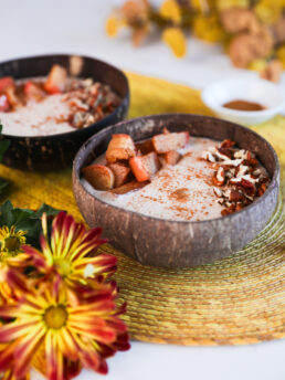 Image of two coconut bowls filled with beige smoothie, garnished with baked apple slices, pecans, and cinnamon, with bright yellow flowers in the fore- and background.