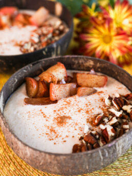 Close up image of two coconut bowls with beige smoothie, garnished with baked apple slices, pecans, and cinnamon, one in focus, the other blurred, with nearby bright yellow flowers.