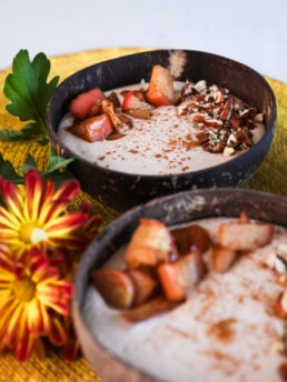 Two coconut bowls with beige smoothie, garnished with baked apple slices, pecans, and cinnamon, one in focus, the other blurred, with nearby bright yellow flowers.