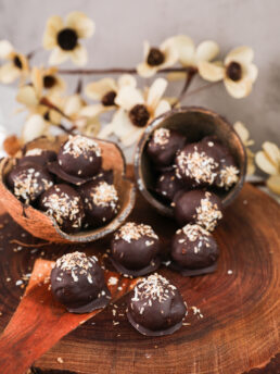 Chocolate-covered balls with a coconut topping displayed in coconut shells, set against a backdrop of beige flowers.