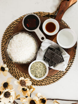 An arrangement of food ingredients, including a bowl of coconut, hemp seeds, maple syrup, and coconut oil, elegantly displayed on a wooden board.
