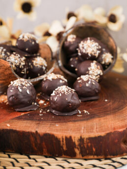 Perspective image of chocolate-covered balls with a coconut topping displayed in coconut shells and sitting on a wooden board, set against a backdrop of beige flowers.