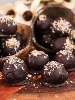 Close up image of chocolate-covered balls with a coconut topping displayed in coconut shells and sitting on a wooden board.