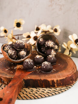 Artistic presentation of dark chocolate-covered balls crowned with shredded coconut, arranged on a wooden board, framed by elegant beige flowers in the background.