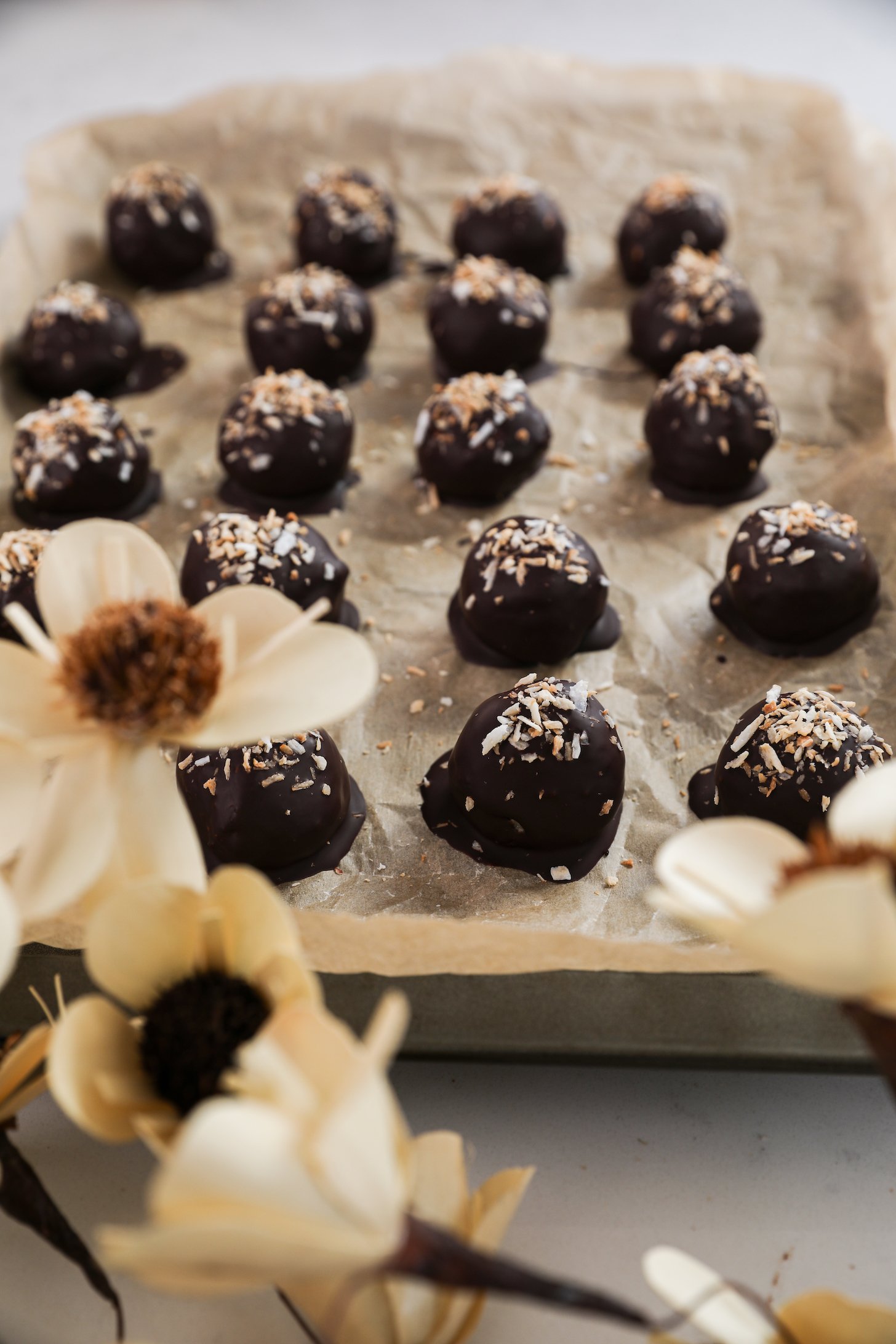 Chocolate-covered balls adorned with shredded coconut, elegantly placed on a parchment paper-lined baking sheet, with delicate flowers in the foreground.