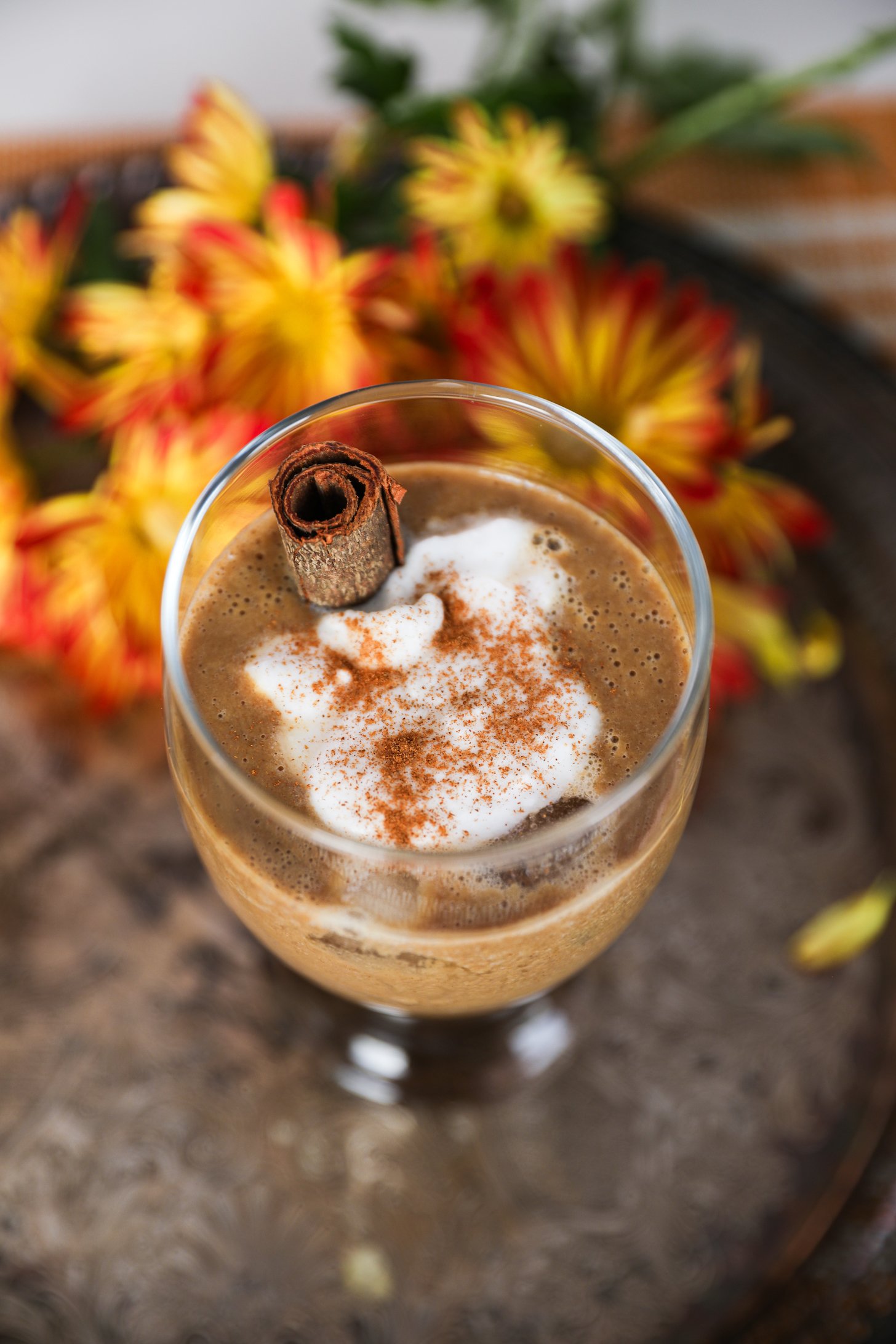 A latte in a glass with cream on top, sprinkled with spice, and a cinnamon stick inside, set against a background of flowers.