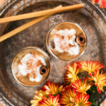 Top-down view of two latte glasses with cream toppings and spice sprinkles, arranged on a brass tray with two straws, accompanied by nearby flowers.