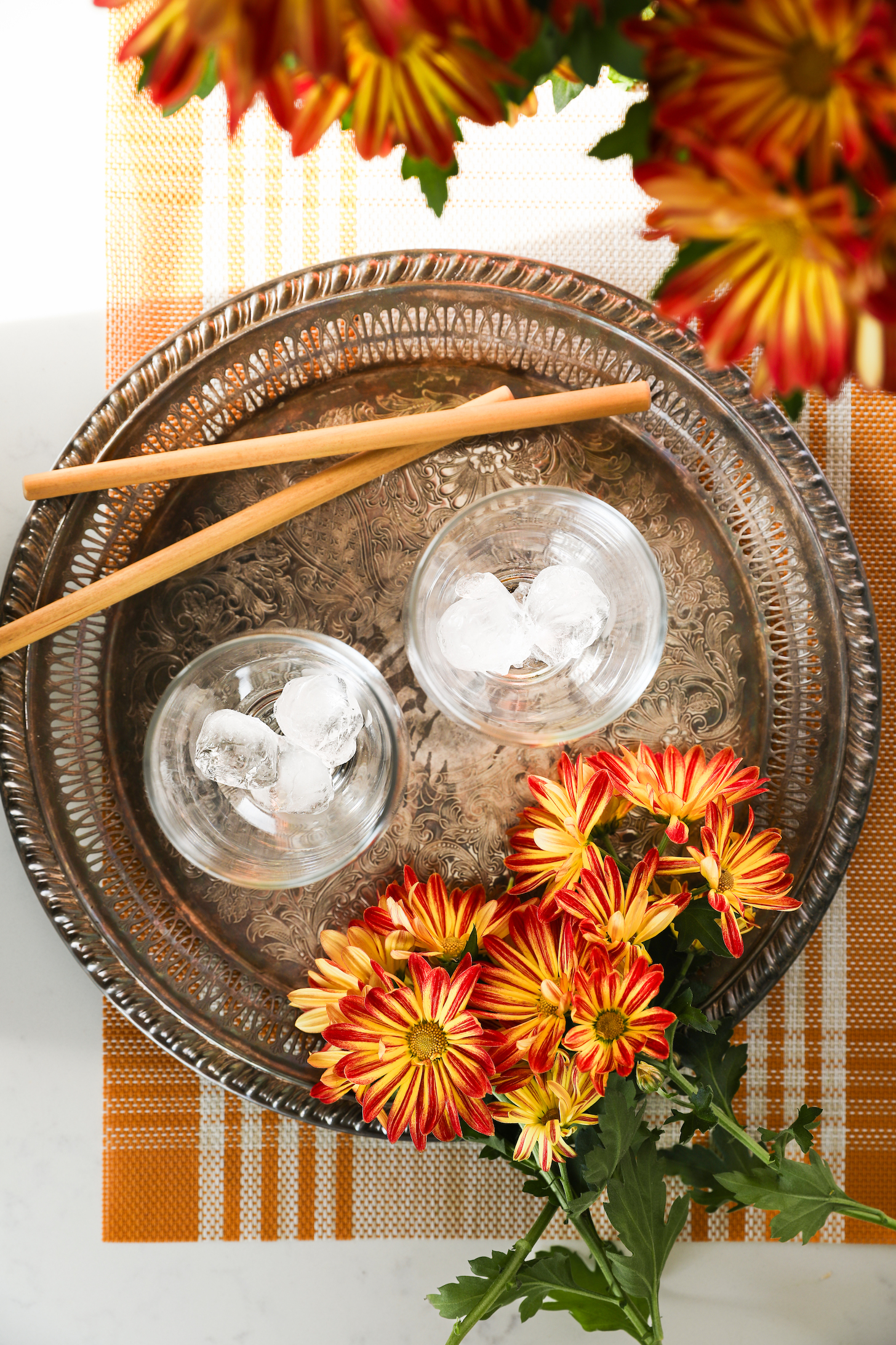 A bird's-eye view of two glasses with ice on a brass tray, accompanied by two straws and nearby flowers.