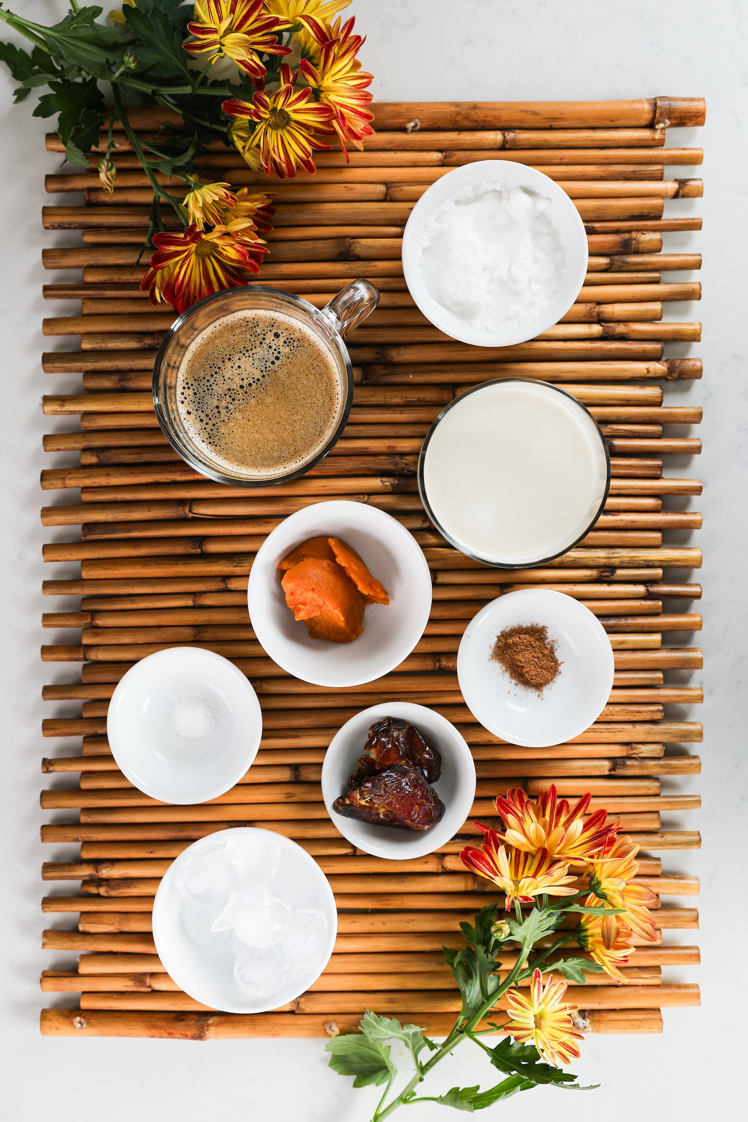A selection of food ingredients, such as milk, coffee, pumpkin puree, dates, and spices, neatly arranged on a bamboo place mat with nearby flowers.