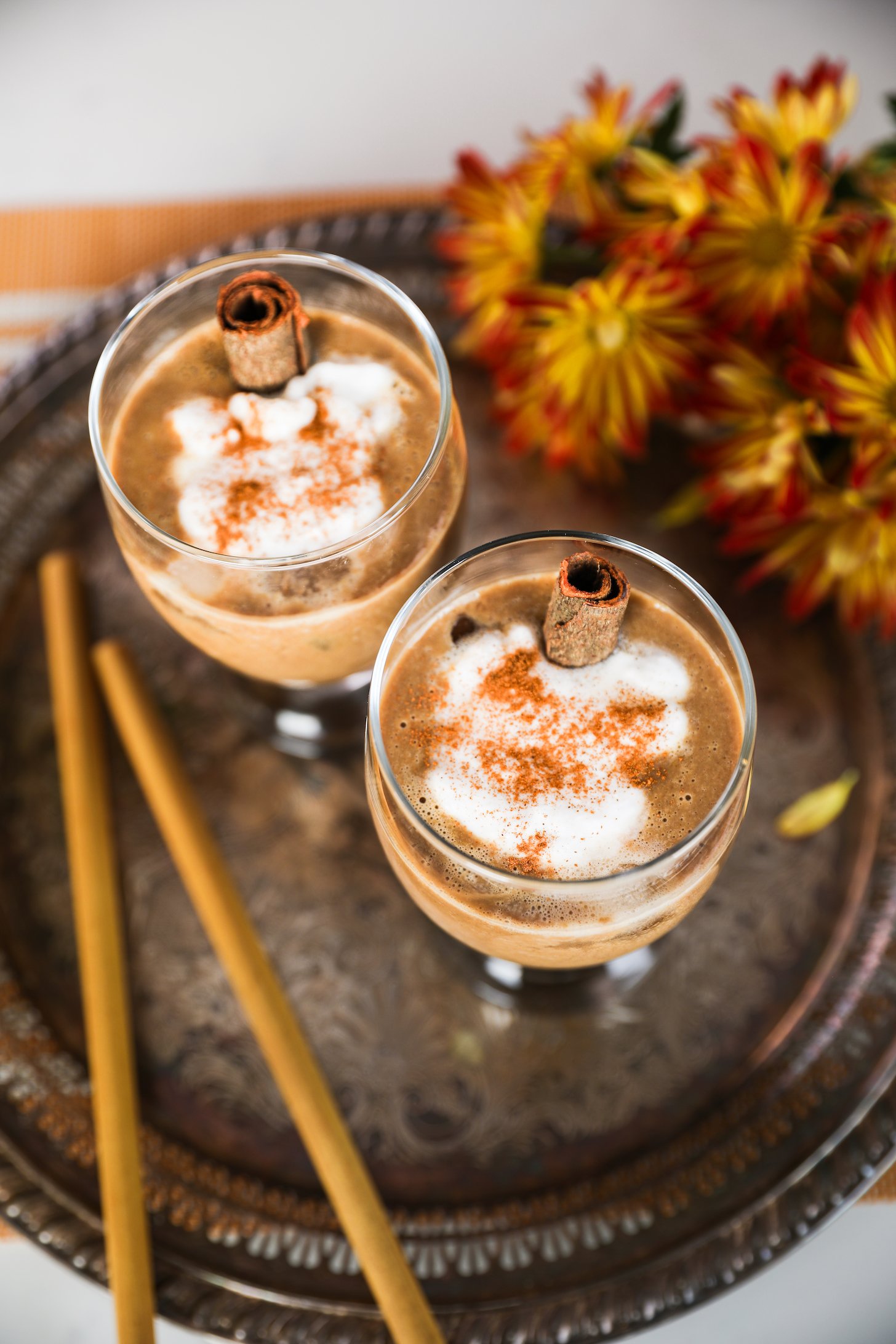 Two latte glasses with cream toppings and spice sprinkles, arranged on a brass tray with two straws, accompanied by nearby flowers.
