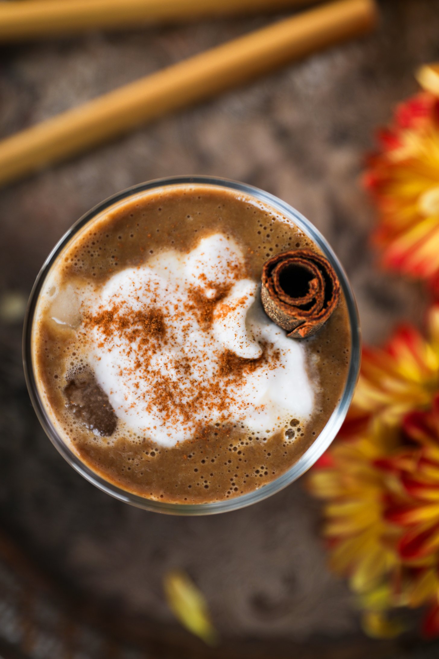 Top-down view of a latte in a glass, with cream on top, sprinkled with spice, and a cinnamon stick inside.
