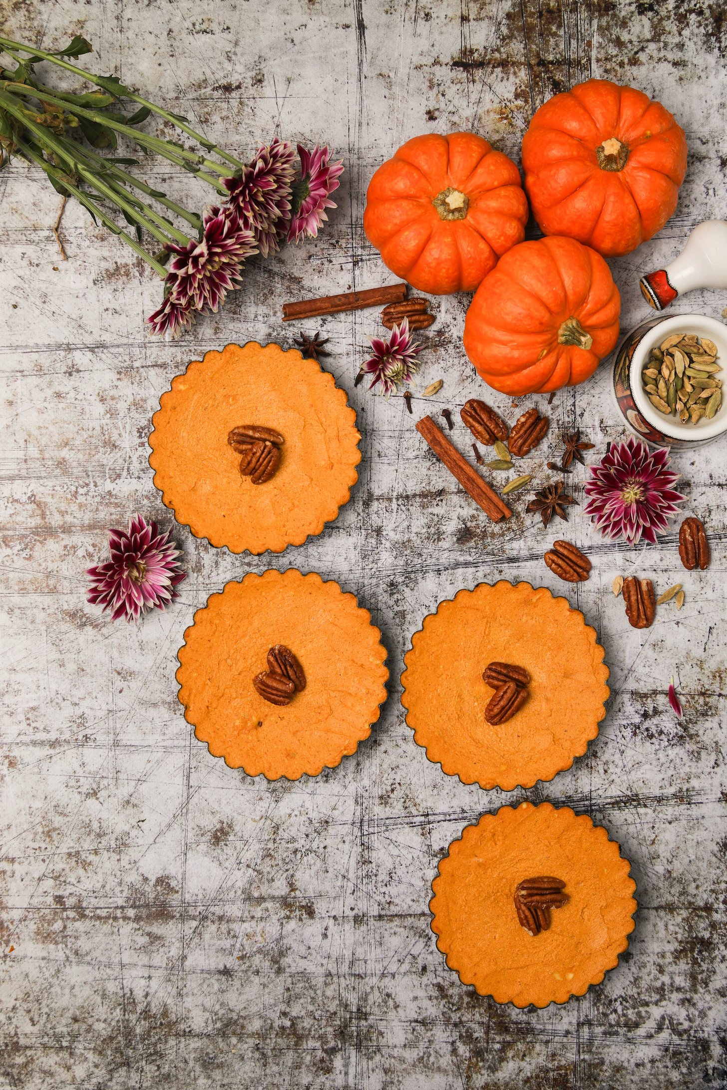Flatlay image of four mini pumpkin pies with pecans in the center, surrounded by mini pumpkins, flowers, spices, and nuts.