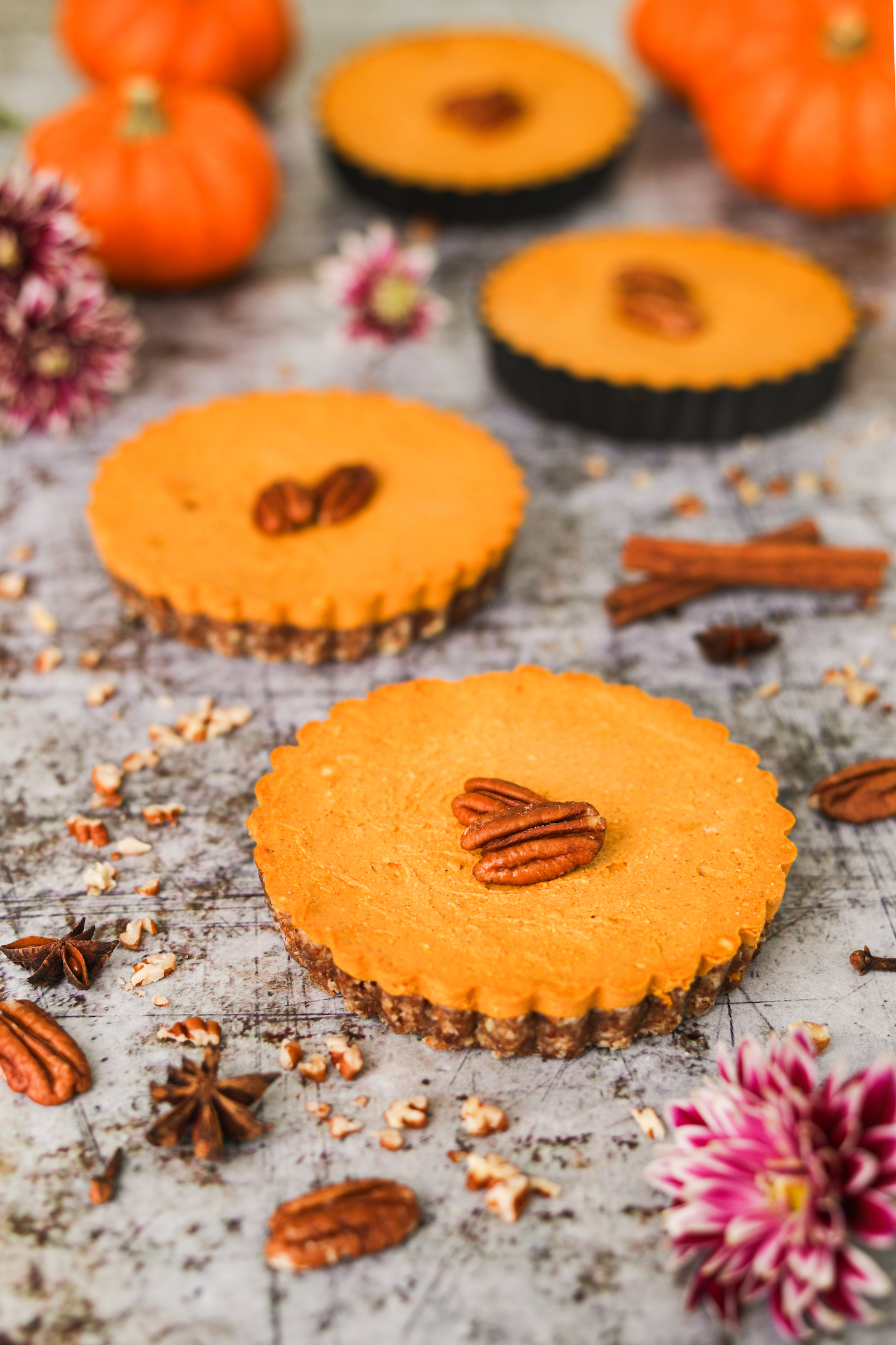 Perspective view of four mini pumpkin pies, each crowned with pecans, artfully arranged amidst flowers, nuts, spices, and whole mini pumpkins.