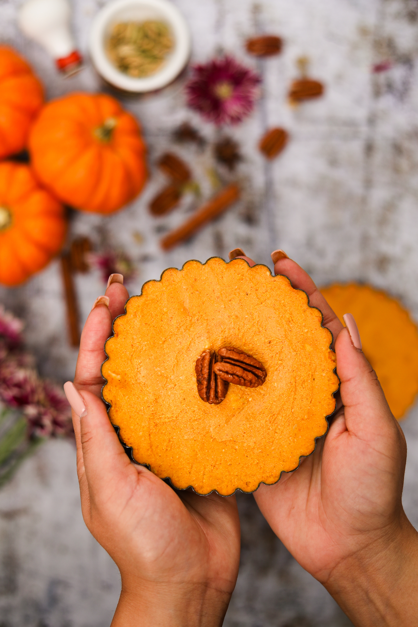 Two hands holding a mini pumpkin pie with two pecans on top, set against a backdrop of mini pumpkins, spices, nuts, and flowers.
