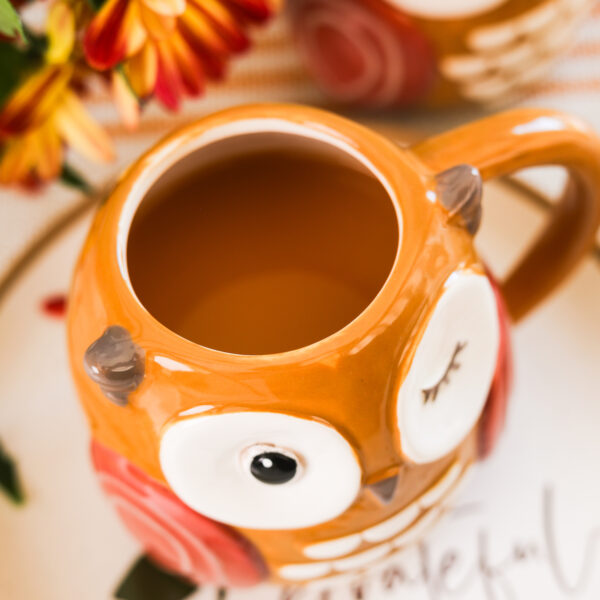 An owl mug filled with chai apple cider rests on a 'grateful' scribed plate against a backdrop of flowers.