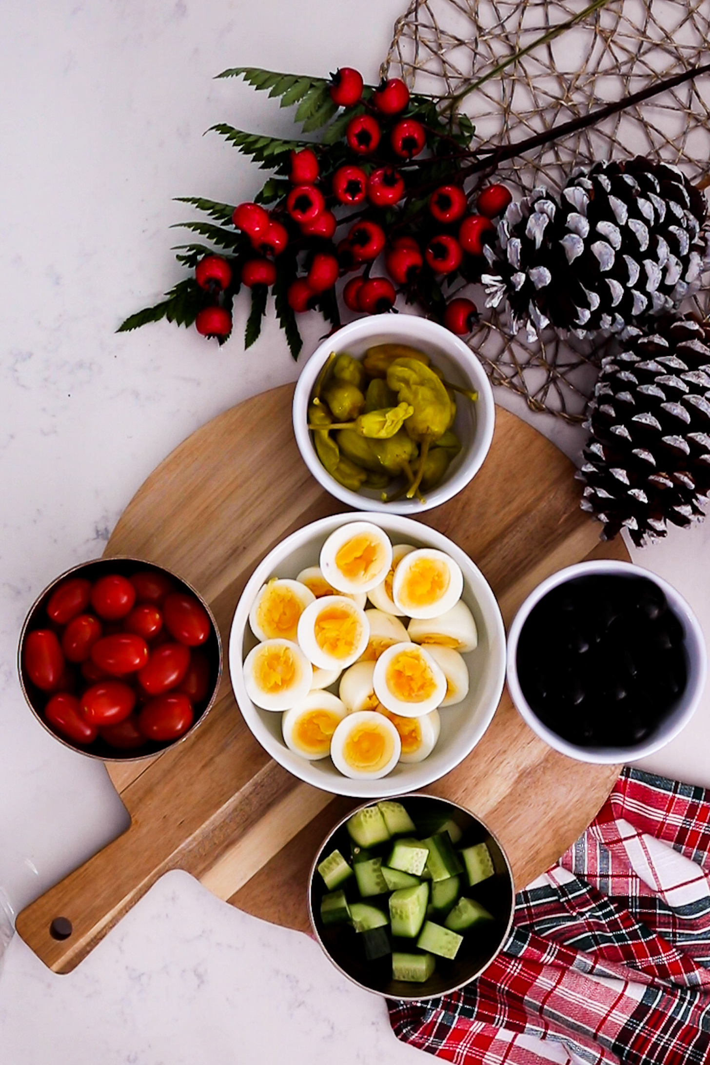 On a wooden board, various food ingredients like boiled egg halves, tomatoes, peppers, cucumber, and olives are arranged in bowls, forming a vibrant collection.