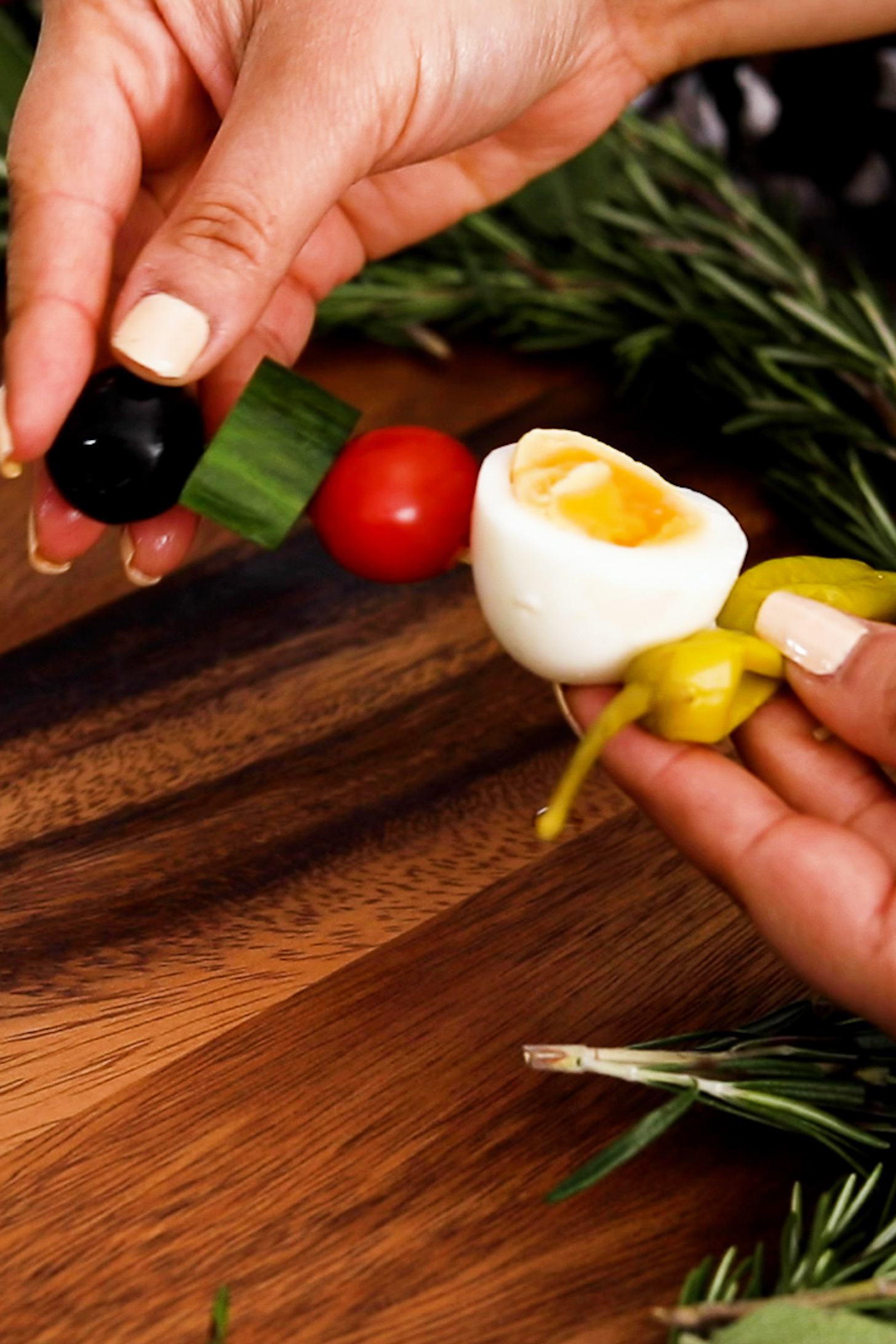 A hand holds a wooden pick threaded with a sequence of pepper, boiled egg, tomato, cucumber, and olive.